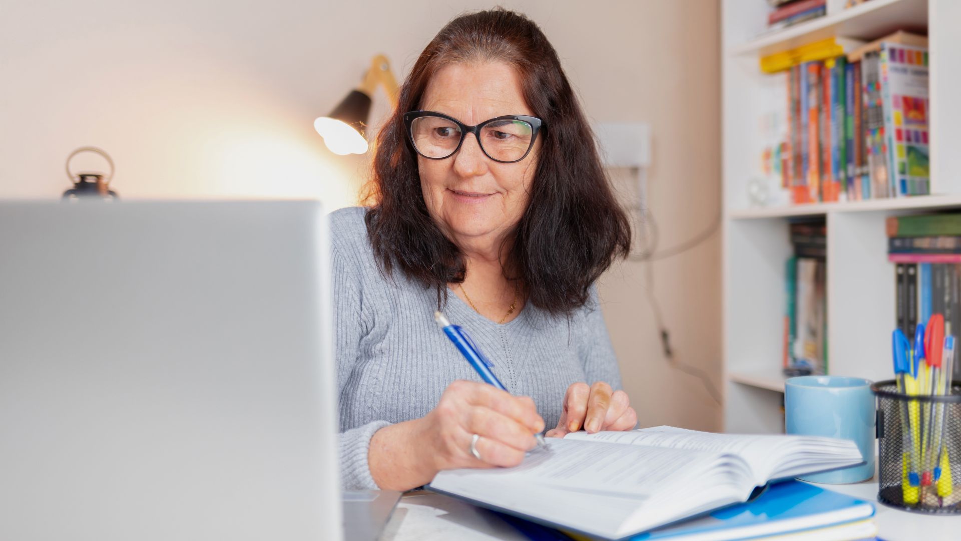 Mulher adulta retomando os estudos pelo EJA da SedeEdu, escrevendo em caderno ao lado do notebook em casa